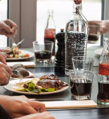 People sharing a meal at a table with dishes of food, including meat and salad. Bottles and glasses of Coca-Cola are present, along with a large bottle of dark liquid. Hands are seen serving and cutting food.