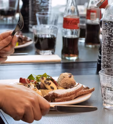A person holds a knife and fork at a dining table. The plate contains lamb chops, potatoes, and vegetables. Soft drink bottles and glasses of water are visible. Other diners are partially seen, enjoying similar meals.