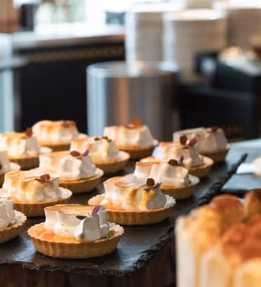 A variety of elegantly arranged pastries and tarts with whipped cream toppings are displayed on a slate board. Some are garnished with small edible flowers. The background shows a blurred scene with stacked plates and kitchenware.