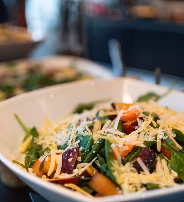 A white bowl filled with a salad containing spinach leaves, diced plums, and a generous topping of grated cheese. The background shows blurred dishes, adding a vibrant, fresh atmosphere to the setting.