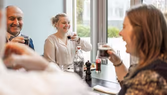 A group of four people are sitting at a table by a large window, smiling and raising glasses in a toast. There are bottles and glasses on the table, and they appear to be enjoying a meal together.