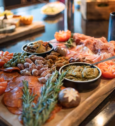 A wooden board displays an assortment of cured meats, sliced tomatoes, olives, nuts, and dip, garnished with rosemary. A glass jar and a black ceramic cup are in the background on a dark countertop.