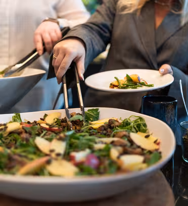 Two people serve themselves a salad from large bowls. The salad includes greens, apple slices, and mushrooms. One person holds a white plate while using tongs. A small jar of seeds and an oil bottle are visible on the side.