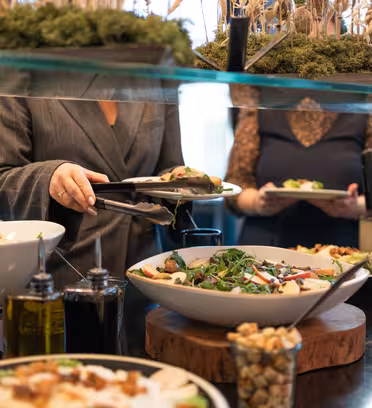 People serving themselves from a buffet with salads in large white bowls. Various toppings and bottles of oil are on the reflective counter. A decorative plant arrangement is above the counter.