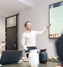 A man in a white shirt is presenting and pointing at a screen with a chart titled Planlægning af processen. A woman with a laptop listens, holding a cup. A meeting room table is visible with drinks and snacks.