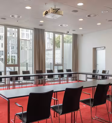 A modern conference room with large windows, red carpet, and minimalistic black chairs arranged around U-shaped tables. The room is well-lit with ceiling lights and equipped with a ceiling-mounted projector.
