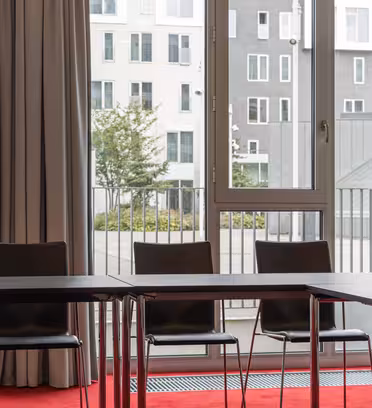 An empty conference room with three black chairs and a long table. Tall windows reveal a view of modern white and gray buildings outside, partially covered by beige curtains. The floor is carpeted in red.