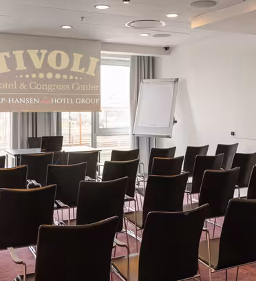 A conference room with rows of empty black chairs facing a projection screen displaying Tivoli Hotel & Congress Center and a flip chart stand. Large windows allow natural light to fill the space.