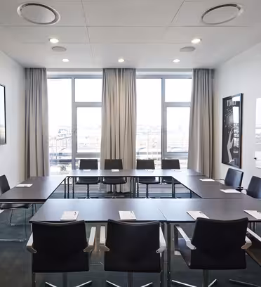 A modern conference room with a U-shaped table arrangement. It has black chairs, a large flat-screen TV on the left wall, a whiteboard on the right, and large windows at the back with beige curtains, allowing natural light in.