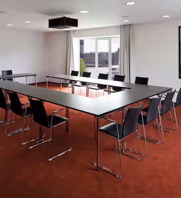 A conference room with red carpet, featuring a U-shaped arrangement of black tables and chairs. A large window with a view outside lets in natural light. A colorful abstract painting hangs on the white wall.