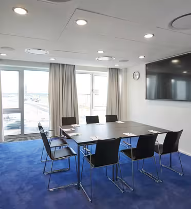 A modern conference room with a large table surrounded by six chairs on a blue carpet. A flat-screen TV is mounted on the wall, and large windows with curtains offer a view outside. Natural light fills the room.