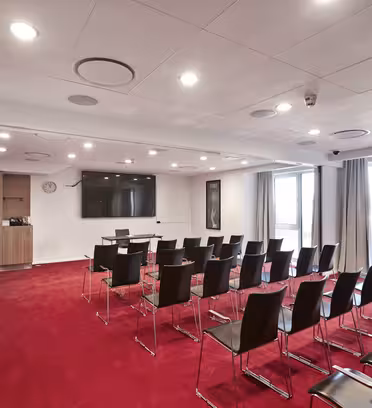 A modern conference room with rows of black chairs on a red carpet facing a wall-mounted flat-screen TV. The room has large windows with beige curtains, bright ceiling lights, and a clock on the wall.
