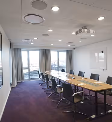 A modern conference room with a long wooden table surrounded by eight black chairs. A projector is mounted on the ceiling, and there is a whiteboard on the wall. Large windows with gray curtains offer a view of the cityscape outside.