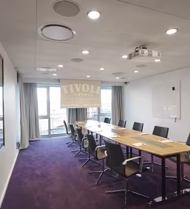 A modern conference room with a long wooden table and ten black chairs arranged around it. The room has a purple carpet, a whiteboard on the wall, and a projector mounted on the ceiling. Large windows at the end provide natural light.