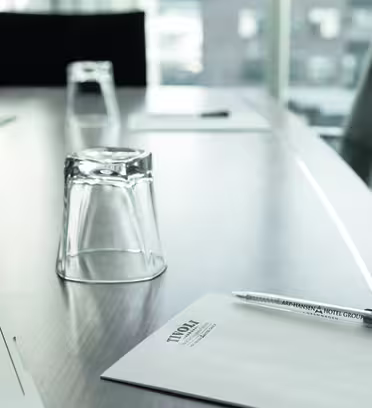 A conference table with a notepad, pen, and an upside-down glass. Blurred office chairs and windows in the background suggest an office setting.