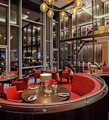 Luxurious restaurant interior with red and black decor. Circular booths and tables are set with wine glasses and napkins. A large wine rack fills the background, and floor-to-ceiling windows offer a cityscape view. Warm lighting illuminates the space.
