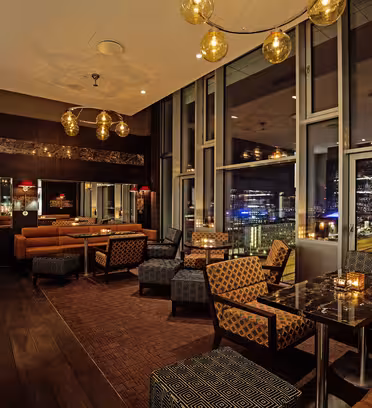 A luxurious, dimly lit lounge with modern décor, featuring leather and patterned chairs, dark wood accents, and floor-to-ceiling windows offering a nighttime city view. Pendant lights with golden globes add a warm ambiance.