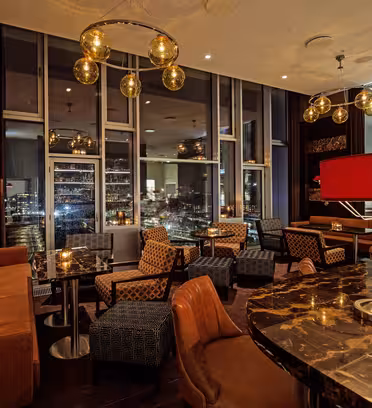 Luxurious lounge with brown leather sofas, patterned chairs, and dark marble tables. Warm lighting from globe pendants and red lampshades. Floor-to-ceiling windows reveal a city skyline at night.