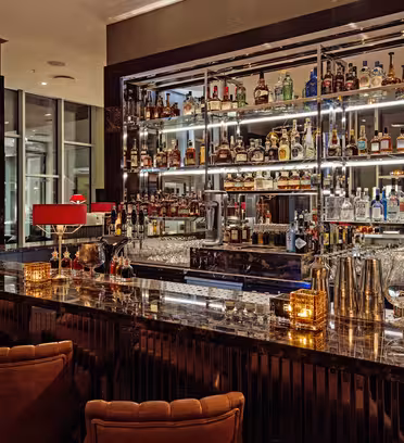 A dimly lit, upscale bar with a polished marble countertop, red lamps, and cushioned stools. Shelves behind the bar are stocked with various bottles of liquor. Glassware and small candle holders adorn the countertop.