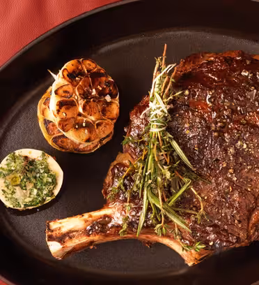 A cooked steak garnished with herbs is on a black oval pan. A grilled half lemon with herbs and a roasted garlic bulb accompany the steak. The pan rests on a red leather surface.