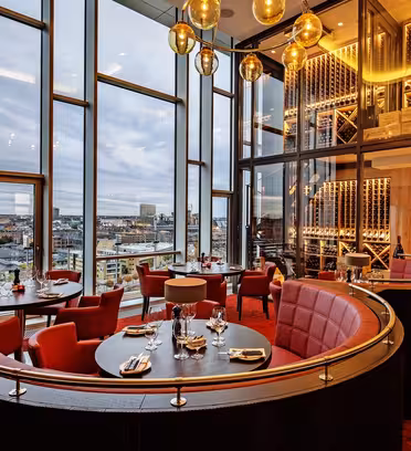 A modern, luxurious restaurant interior with large windows showcasing a cityscape. The space is adorned with red chairs, round tables set for dining, wine racks, and warm lighting from elegant pendant lights.