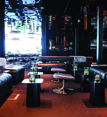 Modern lounge with black leather seating and small tables arranged symmetrically. The space features a glossy ceiling and shelves stocked with glassware. Large windows in the background provide natural light, illuminating the plush, contemporary decor.