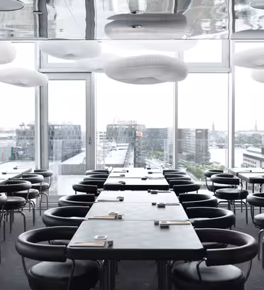 Modern dining area with several tables and round chairs. Large windows provide a view of an urban cityscape. Contemporary light fixtures hang from the ceiling, and each table is set with placemats and utensils.