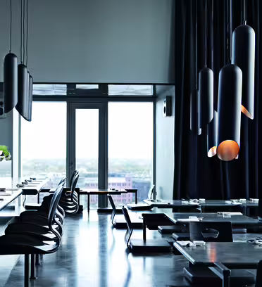 Chic, modern restaurant interior with sleek black tables and unique pendant lights. Large windows provide a view of the cityscape, and black chairs are neatly arranged around the tables, creating an elegant and sophisticated ambiance.