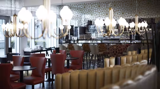 A luxurious, modern restaurant interior with elegant chandeliers, red and beige leather seating, dark wooden tables, and a chic bar area. The wallpaper features a geometric pattern, adding a stylish touch to the welcoming space.