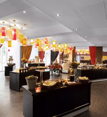 Spacious room with a buffet setup, featuring various food stations with baskets and platters. Colorful ceiling lamps and large windows add brightness. Red and orange curtains accent the space, while wooden floors create a warm atmosphere.