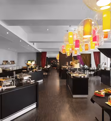 A spacious dining area with a buffet setup. Long tables hold various dishes, and a chocolate fountain is at the forefront. Colorful lamps hang from the ceiling, and curtains in red, pink, and black add a vibrant touch to the room.