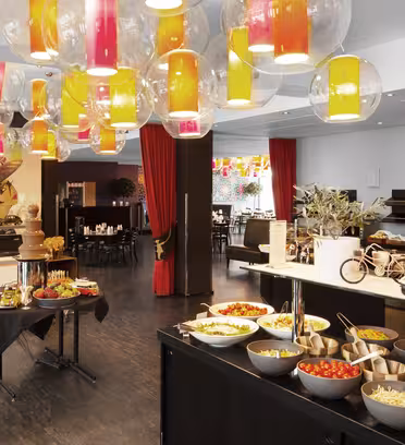 A buffet setup in a brightly decorated room with colorful hanging lights. Various salads, fruits, and snacks are displayed in bowls. Red curtains and wall art add to the lively atmosphere. A chocolate fountain and toy vehicles are also present.