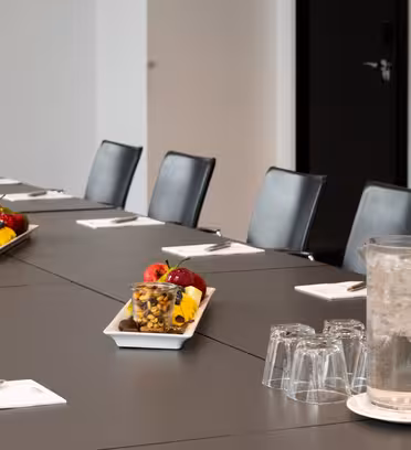 A conference room with a long table, black chairs, and white notepads with pens. The table has trays with fruit and nuts, and a pitcher of water with empty glasses. A door and cabinet are in the background.