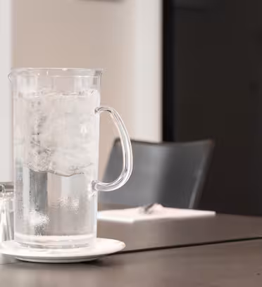 A clear glass pitcher filled with ice water sits on a small white plate on a dark table. Four upside-down glasses are nearby. The background features chairs and an open door in a minimalist room setting.