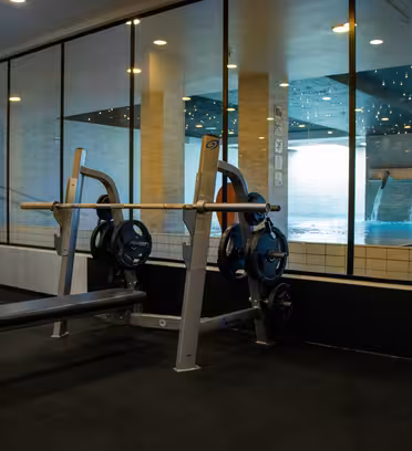 A gym bench press station with weights is placed near a large window. Outside, there is a view of a pool with water spouts and lounge chairs. The room has a modern design with black flooring and white walls.