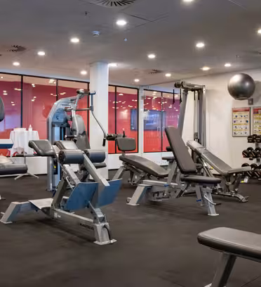 A modern gym with various exercise equipment including weight machines, benches, and dumbbells. The room features large windows, bright lighting, and fitness posters on the walls. The floor is covered with black mats.