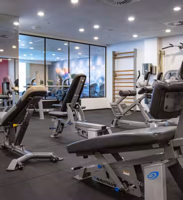 A modern gym featuring various fitness equipment, including benches and weight machines. The room has a mirrored wall, glass partitions, and a wooden ladder for exercises. The atmosphere is clean and spacious with dark flooring.