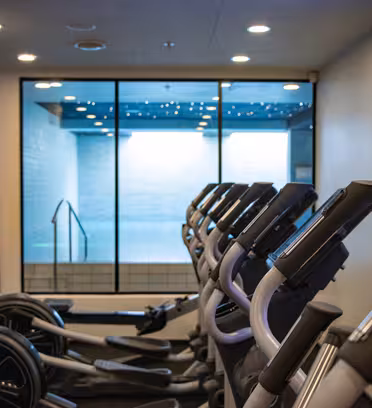 Row of stationary exercise bikes in a gym facing a wall-mounted TV. Large window reveals a view into an indoor pool area with a modern ceiling.