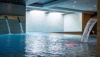 Indoor swimming pool with two cascading water features on the left and one large water spout on the right. The ceiling is dark, enhancing the bright natural light coming from the windows on the left wall, creating a serene atmosphere.