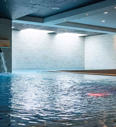 Indoor swimming pool with two cascading water features on the left and one large water spout on the right. The ceiling is dark, enhancing the bright natural light coming from the windows on the left wall, creating a serene atmosphere.