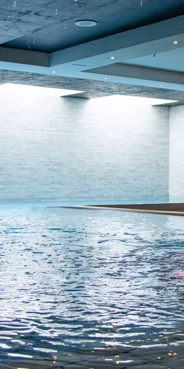 Indoor swimming pool with two cascading water features on the left and one large water spout on the right. The ceiling is dark, enhancing the bright natural light coming from the windows on the left wall, creating a serene atmosphere.