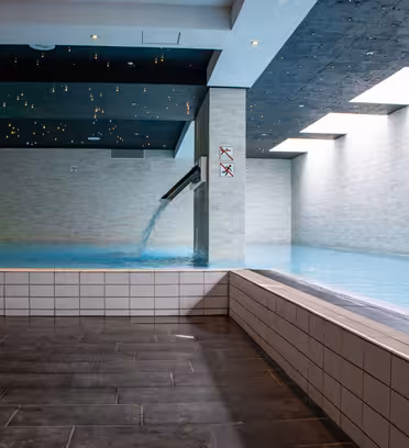 Indoor pool area with brick walls and a rectangular pool. Water flows from a spout at the corner. The ceiling has small star-like lights, and large windows let in natural light. The floor is tiled in dark gray.