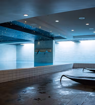 Indoor swimming pool area with a modern design. The pool is illuminated with soft blue lights and surrounded by tiled walls. Two sleek, dark lounge chairs are on the wet floor, and the ceiling is dotted with small lights.