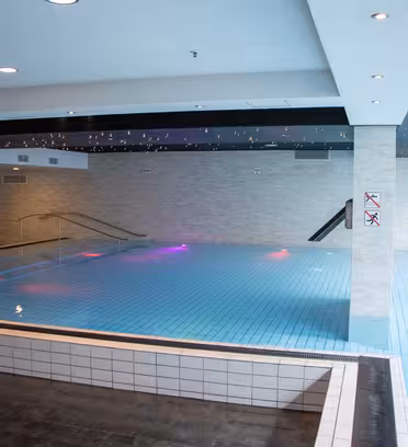 Indoor swimming pool with clear blue water and tiled floor. Soft lighting creates a calm ambiance. Steps lead into the pool, and a railing provides support. No diving signs are visible on the wall. Ceiling has a pattern resembling a starry night.