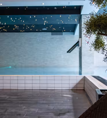 Indoor swimming pool with a sleek, modern design. The ceiling has star-like lighting, and a potted plant is in the foreground. The pool area features a tiled floor and minimalistic decor, creating a serene atmosphere.