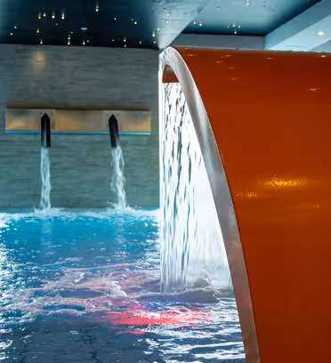 Indoor swimming pool with modern water features. A curved orange structure pours water into the pool, and two wall-mounted spouts contribute streams as well. The pool area is illuminated, creating a serene atmosphere.