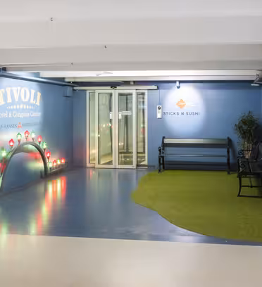 Entryway to a hotel and sushi restaurant decorated with a small artificial turf area, benches, and colorful lights shaped like mushrooms. Blue walls feature logos for Tivoli Hotel & Congress Center and SticksnSushi.