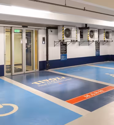 Underground parking area with blue accessible parking spaces marked on the floor. Sign for Tivoli Hotel & Congress Center displayed. Two wall-mounted ventilation units and an entrance door are visible.