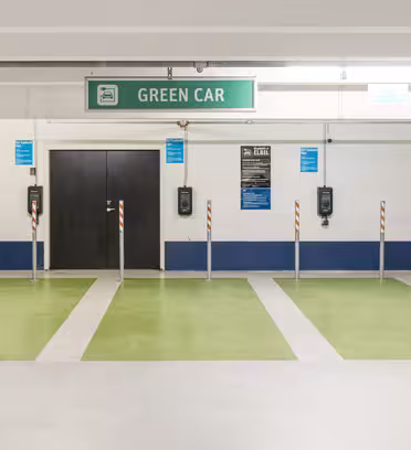 A parking garage featuring multiple green electric vehicle charging spots with a prominent Green Car sign above. Black chargers are mounted on the wall, and the area is marked by bright green paint on the floor.