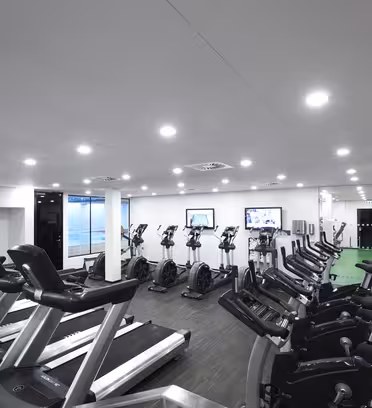 A modern gym with multiple treadmills and stationary bikes lined up in rows. The space is well-lit with ceiling lights, and there are two wall-mounted TVs at the far end. The floor is dark, contrasting with the white walls.